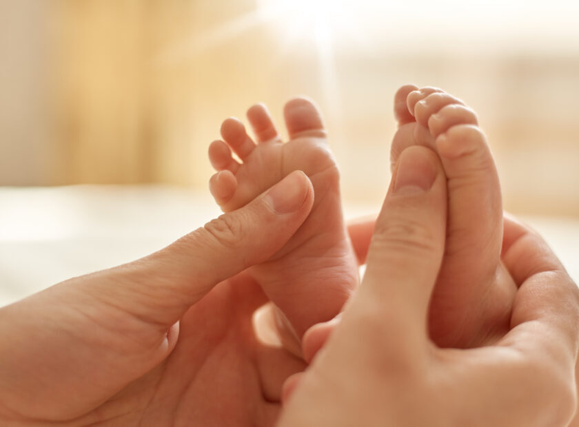 Mum making baby massage, mother massaging infant bare foot, preventive massage for newborn, mommy stroking the baby's feet with both hands on light background.