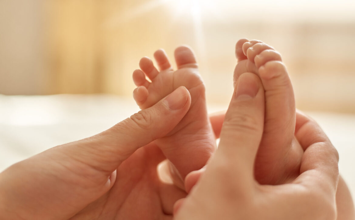 Mum making baby massage, mother massaging infant bare foot, preventive massage for newborn, mommy stroking the baby's feet with both hands on light background.